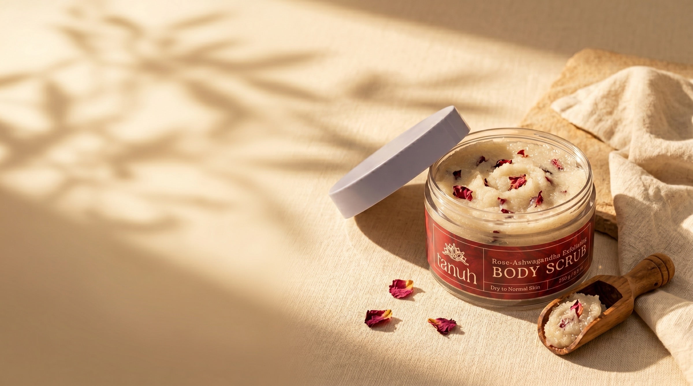Tanuh Rose & Ashwagandha Body Scrub jar, open with a wooden scoop and dried rose petals, on cream linen in warm natural light